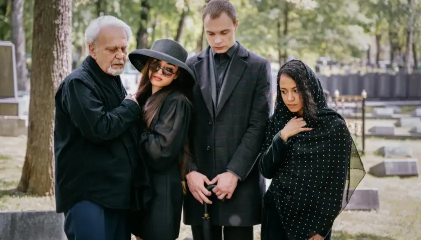 Smutna rodzina na cmentarzu. Elegancki ubiór damski na pogrzebie zimą, z kapeluszem i okularami przeciwsłonecznymi, obok żałobników w czerni.