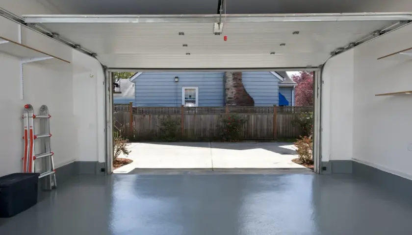 Interno di un garage con porta sezionale aperta su un cortile. Si vede una scala e un contenitore. Pensare a quanto costa trasformare un garage in abitazione.