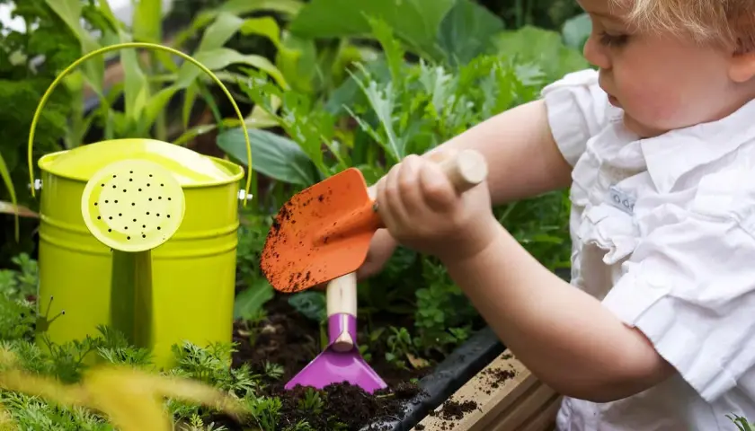 Kleines Kind mit Schaufel im Garten, lernt, wie man ein kinder hochbeet selber baut. Grüne Gießkanne daneben.