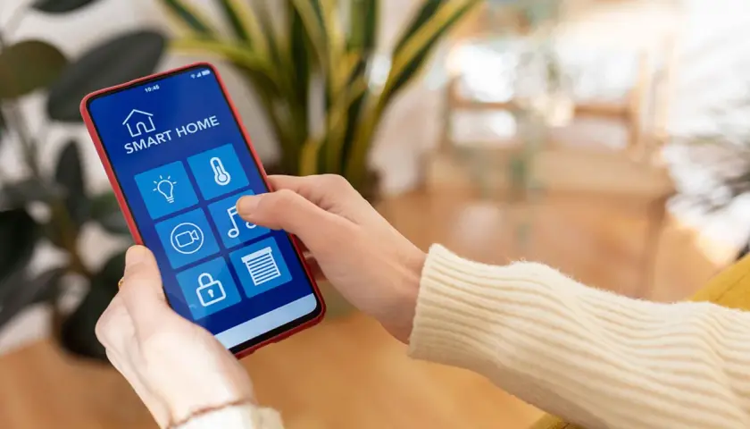 Mano usando un smartphone para controlar productos inteligentes de hogar: luces, temperatura, cámara, música y persianas.