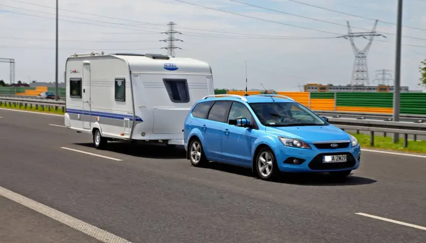 Niebieski Ford Focus z przyczepą kempingową Hobby na drodze. Zestaw mieści się w limitach maksymalna długość pojazdu z przyczepą kat b.