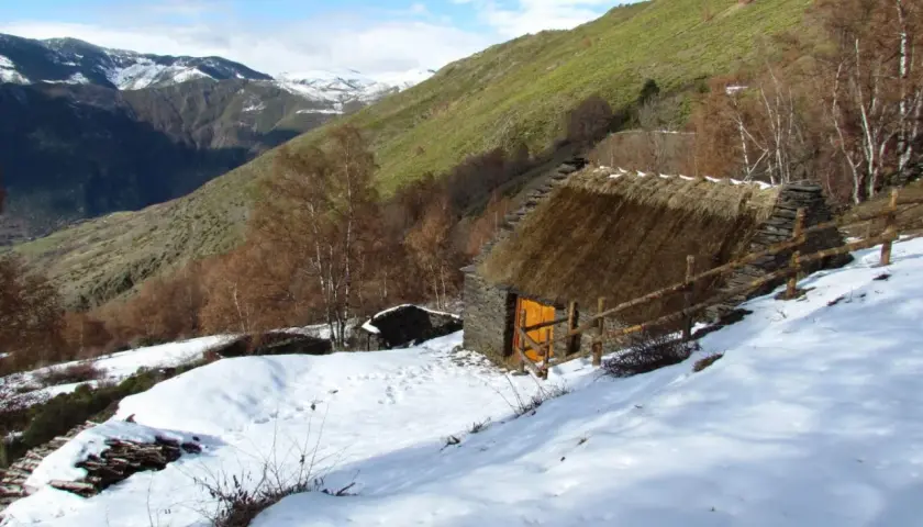 Pequeña cabaña de piedra con tejado de paja, rodeada de nieve y un paisaje montañoso. Un esterri de cardos se alza en la ladera verde.