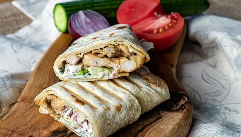 Pyszny kebab w tortilli z kurczakiem, warzywami i sosem, podany na desce z pomidorem i ogórkiem.