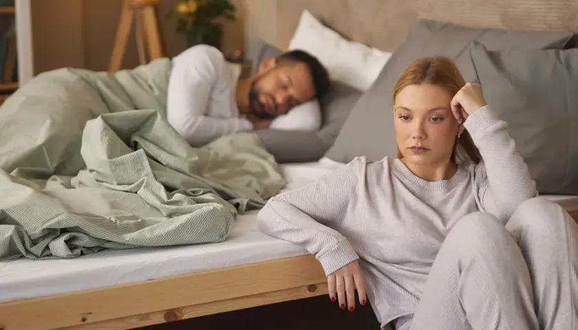 Eine Frau sitzt nachdenklich am Bettrand, während ihr Partner schläft. Sie kämpft mit ihrer Sexsucht bei Frauen.