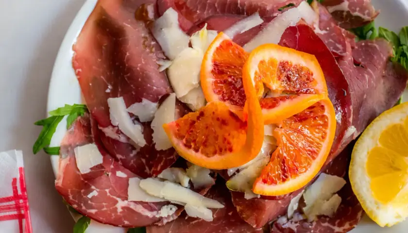Wykwintna szynka bresaola z płatkami parmezanu, krwistymi pomarańczami i cytryną na talerzu.