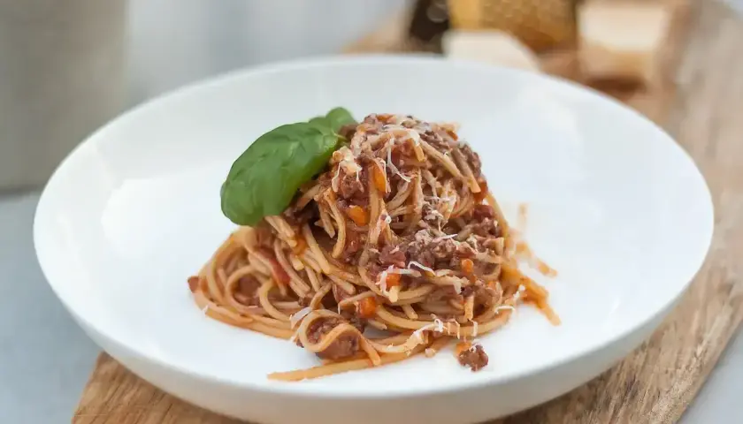 Porcja spaghetti bolognese z parmezanem i listkiem bazylii na białym talerzu.