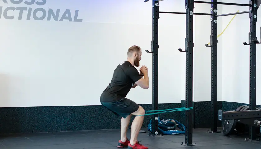 Mężczyzna wykonuje ćwiczenie spanish squat z gumą oporową wokół ud, w siłowni.