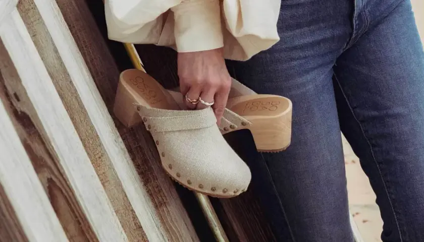 Une femme tient un sabot femme en toile beige, orné de clous, avec un talon en bois. Elle porte un jean bleu.