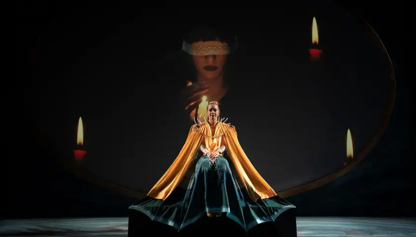 Scena teatrale ispirata al circe film: una donna con mantello dorato, circondata da candele, sotto un'immagine proiettata di una figura bendata.