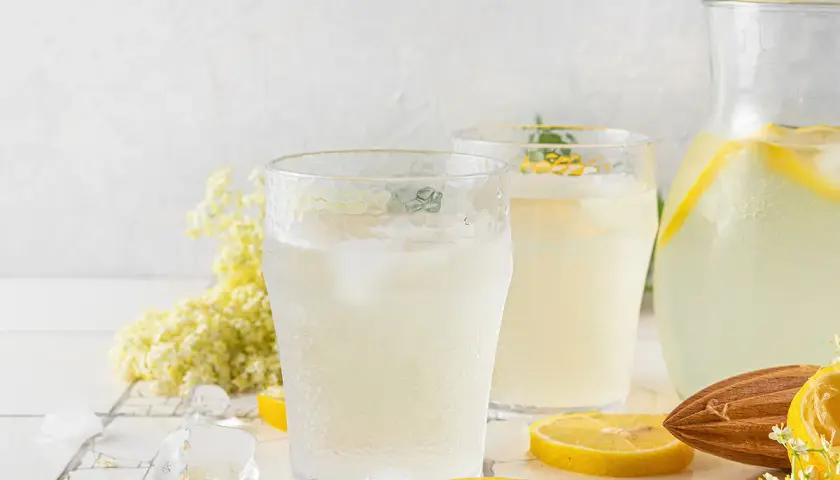 Deux verres de citronnade maison rafraîchissante avec des glaçons, des rondelles de citron, des fleurs de sureau et une carafe remplie.