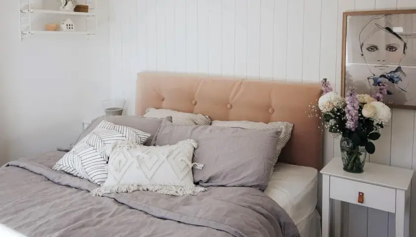 A cozy bedroom featuring a DIY upholstered headboard in a soft peach velvet, adorned with tufted buttons. The bed is dressed in linen bedding and an assortment of decorative pillows.