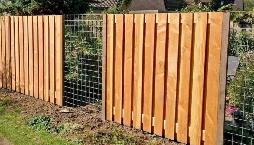 Clôture en bois reconstitué, avec des planches verticales et une section grillagée. Un chien est visible derrière le grillage.