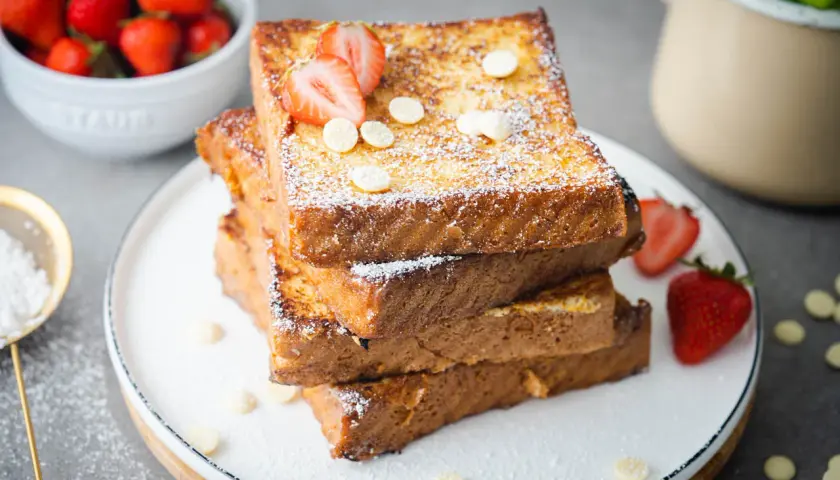 Gestapelte French Toast Scheiben, bestäubt mit Puderzucker und garniert mit Erdbeeren und weißen Schokoladenchips.