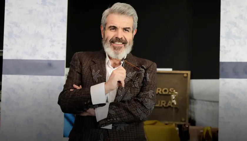 Lorenzo Caprile, con su característica barba canosa, sonríe mientras sostiene unas gafas. Viste una chaqueta de tweed con bordados florales.