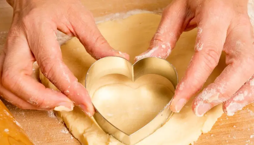 Manos cortando masa con un cortador en forma de corazón, parte del proceso de como hacer galletas caseras.