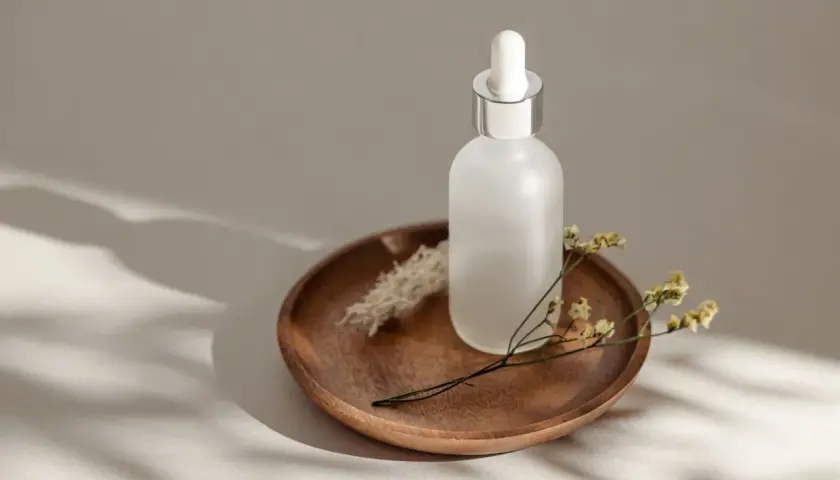 Szklana buteleczka z pipetką na drewnianej tacy, ozdobiona suchymi kwiatami. Uosobienie slow beauty, spokoju i naturalności.