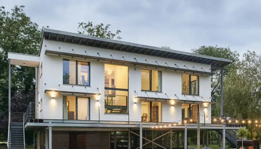 Modernes Haus auf Stelzen mit großen Fenstern und Metalltreppen, umgeben von Bäumen. Lichterketten beleuchten die Terrasse.