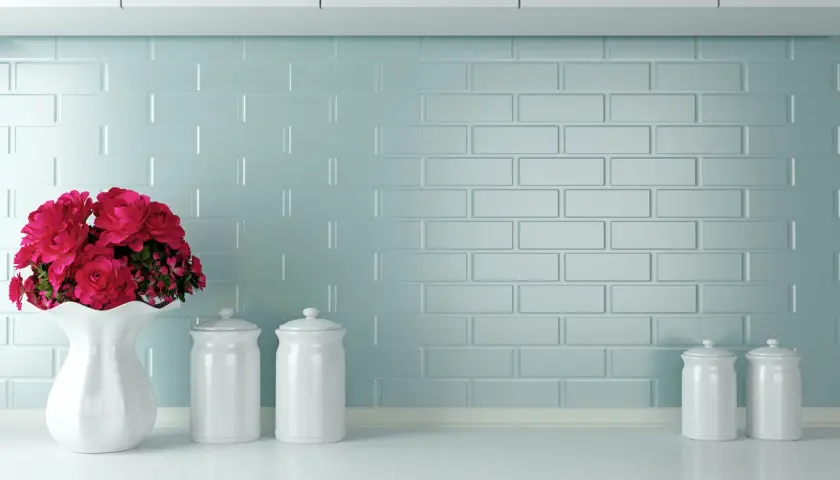 Un mazzo di rose rosse in un vaso bianco su un piano di lavoro, con barattoli bianchi e uno sfondo di piastrelle azzurre. Ottime opinioni per dipingere piastrelle cucina.
