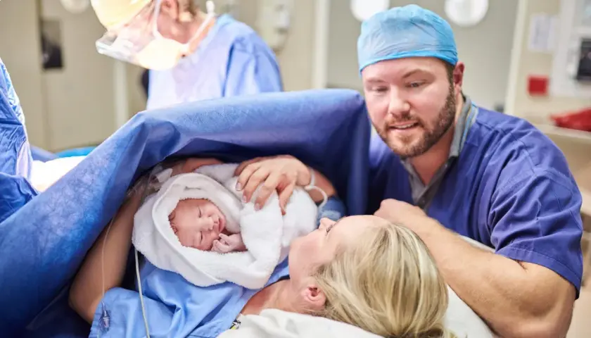 Un padre guarda con amore il suo neonato, avvolto in un asciugamano bianco, dopo un cesareo dolce. La madre sorride, circondata da personale medico.