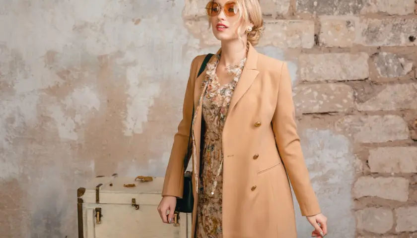 Blondynka w płaszczu w kolorze camel, idealnym dla niej. W tle stara walizka i ceglana ściana.