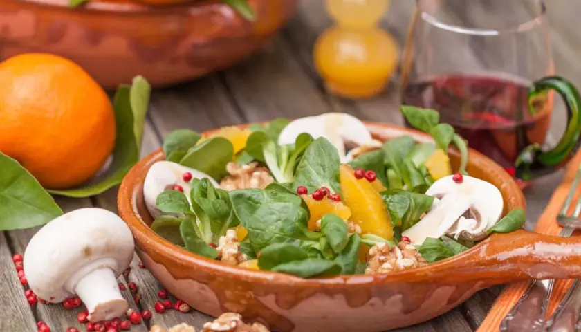 Ein frischer Wintersalat mit Orangen, Pilzen und Walnüssen in einer Terrakotta-Schale. Daneben liegen frische Orangen und ein Glas Rotwein.