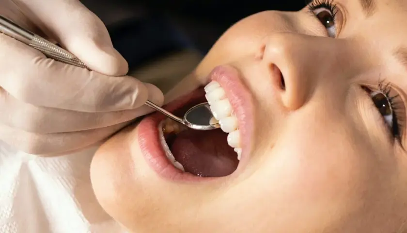 Uśmiechnięta pacjentka podczas zabiegu plastyka dziąseł. Dentysta bada zęby lusterkiem. Efekt plastyka dziąseł przed i po jest widoczny.