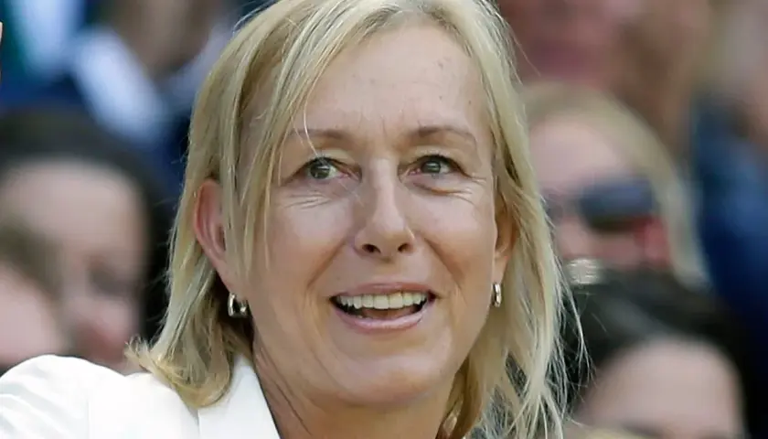 Uśmiechnięta Martina Navratilova w białej marynarce, z blond włosami i kolczykami w kształcie kółek, obserwuje wydarzenie.