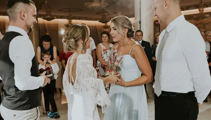 Nietypowe podziękowania dla rodziców na weselu: panna młoda w koronkowej sukni wręcza bukiet w szkle, a pan młody prezent z wstążką.