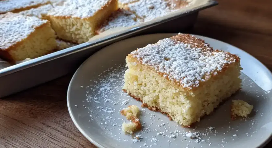 En bit fluffig, nybakad mjuk kaka med pudersocker på en grå tallrik. Resten av kakan syns i en ugnsform i bakgrunden.