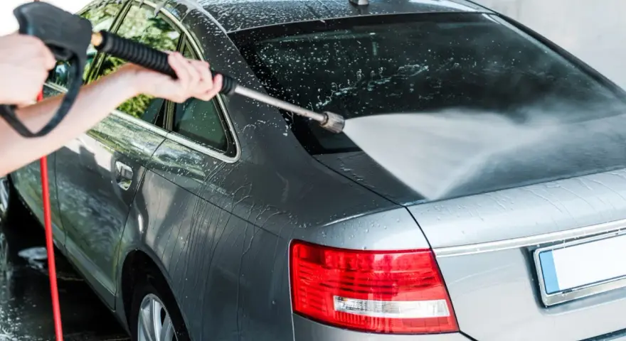 A person demonstrates how to power wash a car, spraying water onto the rear of a grey sedan.