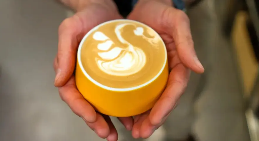 Dłonie trzymają żółtą filiżankę z kawą, na której widnieje wzór łabędzia. Zastanawiasz się, ile kofeiny ma espresso?