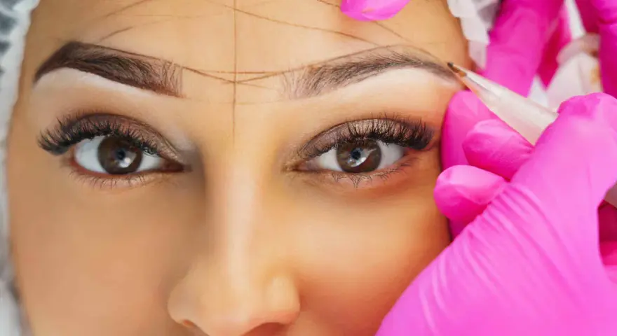 Nahaufnahme einer Frau, deren Augenbrauen mit Linien markiert werden, während eine Person in pinken Handschuhen mit einem Stift arbeitet.