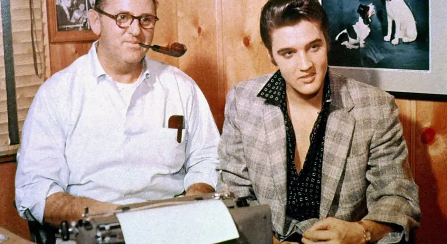 Tom Parker und Elvis Presley sitzen nebeneinander. Parker, mit einer Pfeife im Mund, tippt auf einer Schreibmaschine.