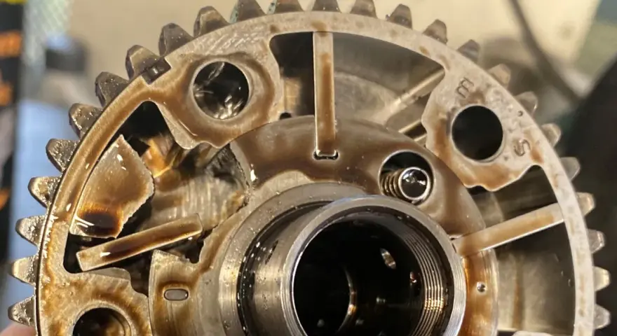 Metalowe koło zmiennych faz rozrządu, pokryte olejem, z widocznymi elementami mechanicznymi i zębatkami.