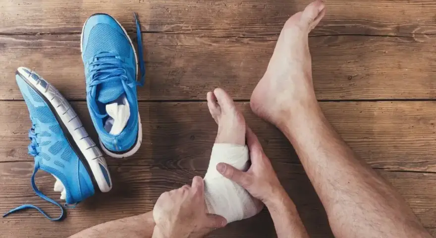Noga z bandażem na kostce, obok niebieskie buty sportowe. Wciąż widoczna opuchlizna 2 miesiące po skręceniu kostki.