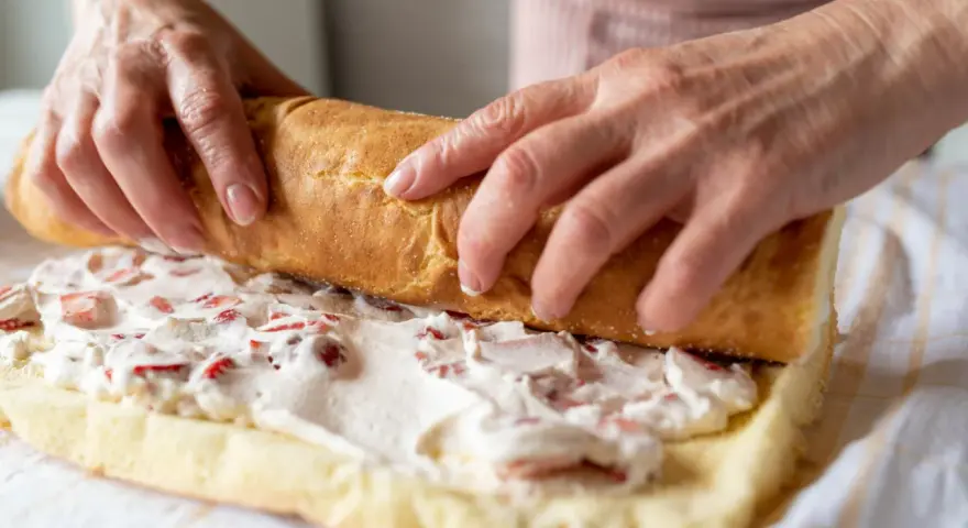 Ręce zwijają biszkopt z kremem i truskawkami. To najprostsza rolada biszkoptowa, która zachwyci smakiem.