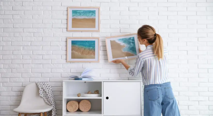 Mujer colocando cuadros de playa en pared de ladrillo blanco. Ideas para como combinar cuadros en la pared.