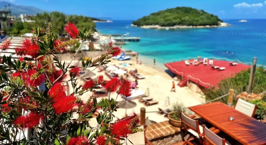 Albania camping nad morzem: czerwone kwiaty na tarasie z widokiem na piaszczystą plażę, turkusowe morze i zieloną wyspę.