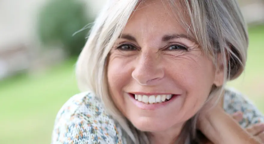 Mujer sonriente con cabello rubio y arrugas normales a los 50 años, disfrutando de un día al aire libre.