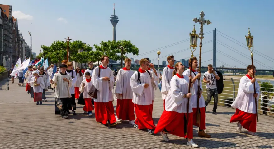 Jugendliche in Soutanen und Alba nehmen an einer Prozession teil, die an christliche Feiertage erinnert. Sie tragen Kreuze und Fahnen.