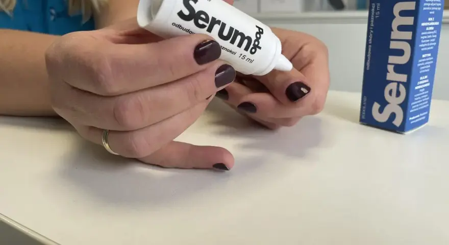 Dłonie z ciemnym lakierem aplikują podoserum na paznokieć. Opakowanie Podo Serum widoczne obok.