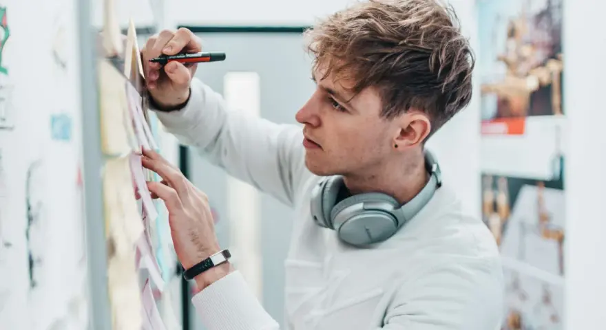 Ein junger Mann mit Kopfhörern am Hals und einem Stift in der Hand, der Ideen für sein Mediendesign Studium an einer Pinnwand sammelt.