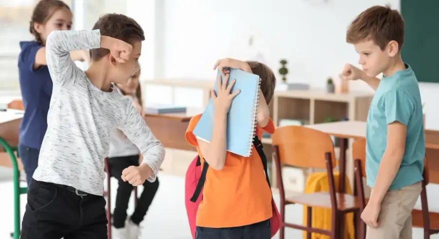 Chłopcy w klasie doświadczają konfliktów między rówieśnikami. Jeden chłopiec grozi pięścią, inny zasłania się książką.