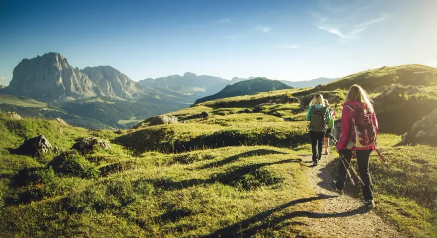 Persone in un percorso di trekking e hiking tra le montagne, con zaini e bastoncini da passeggio, sotto un cielo azzurro.