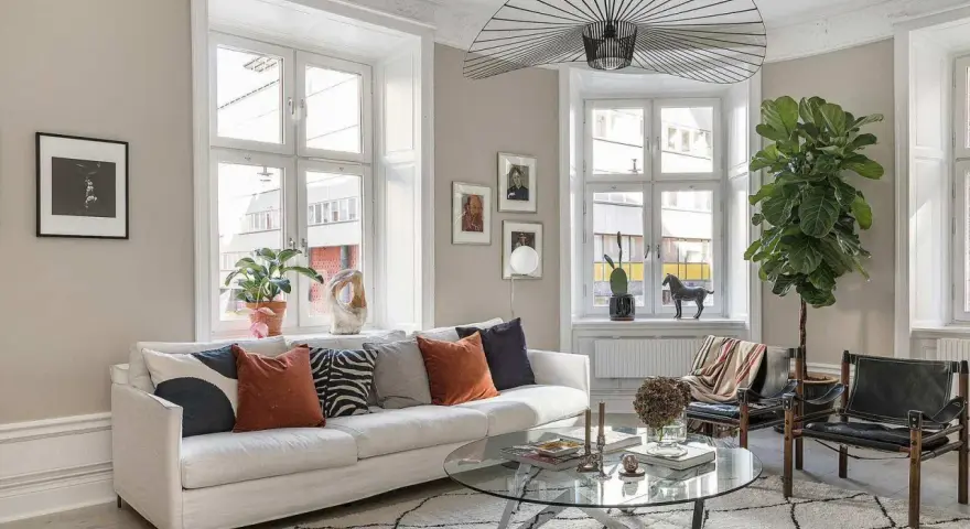 Vardagsrummet är ljust med en vit soffa, kuddar i olika färger och mönster, ett runt soffbord och en stor taklampa. En stor grön växt bidrar till trivseln.