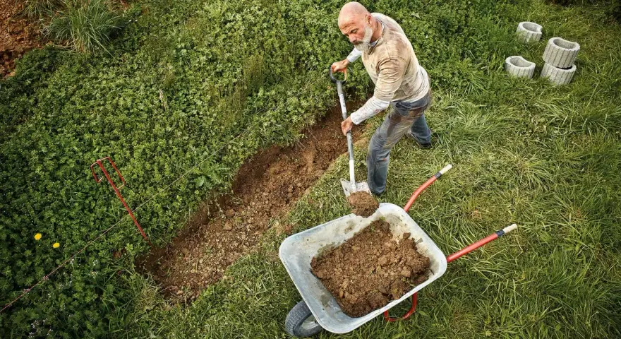 Mann gräbt einen Graben, um den Hang abzufangen. Erde wird in eine Schubkarre geschaufelt.