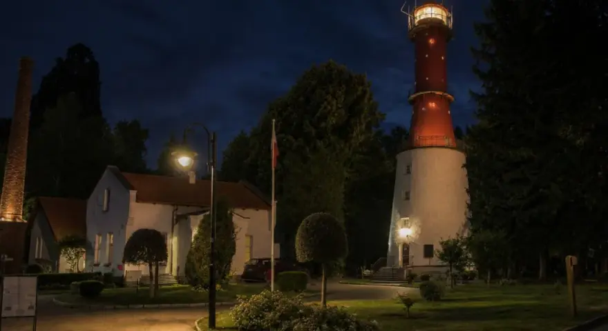 Nocą rozświetlona latarnia morska Rozewie strzeże wybrzeża. Obok niej stoją budynki i ceglany komin.