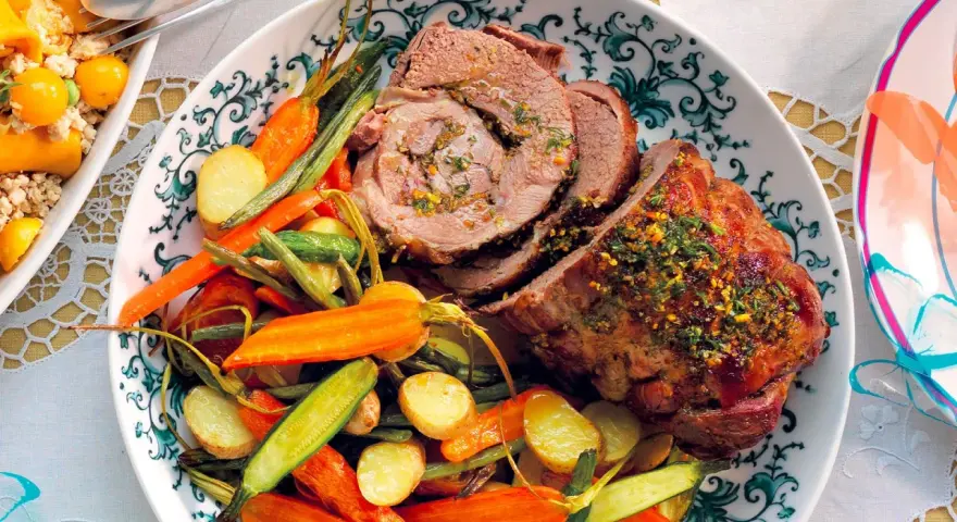 Agnello al forno succulento, affettato e servito con patate novelle, carote e fagiolini croccanti su un piatto decorato.