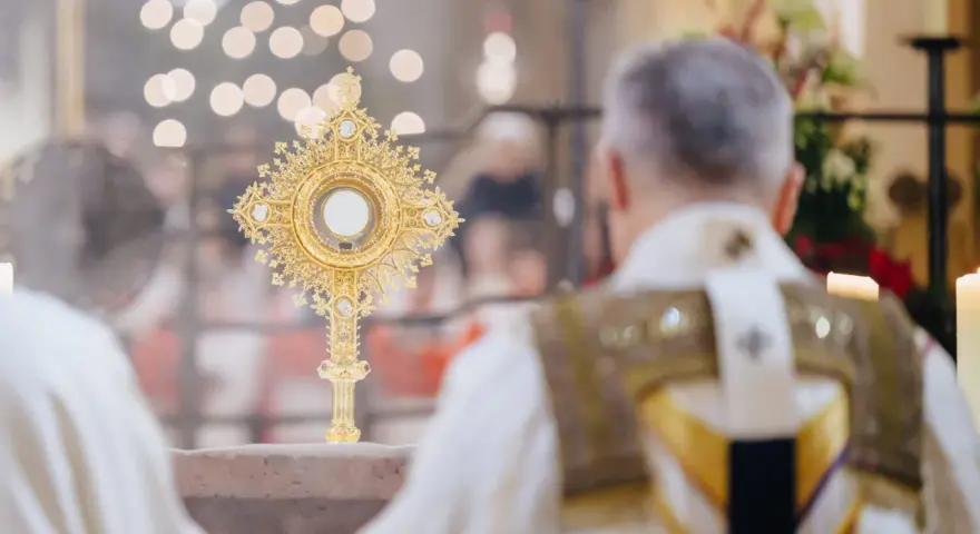Goldene Monstranz mit Hostie im Schein von Kerzenlicht. Ein Priester in festlicher Kleidung feiert die Messe, ein Moment des weihnachtlichen Christentums.