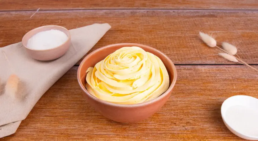 Una ciotola piena di soffice crema al burro, con zucchero in due piccole ciotole e decorazioni di erba secca su un tavolo di legno.
